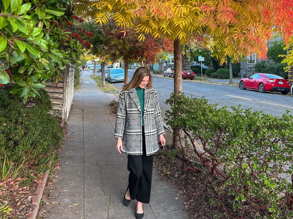 A young, light-haired woman wears a green sweater, black and white houndstooth coat, and black pants while walking towards the camera on a residential sidewalk, with yellow autumn leaves surrounding her. 