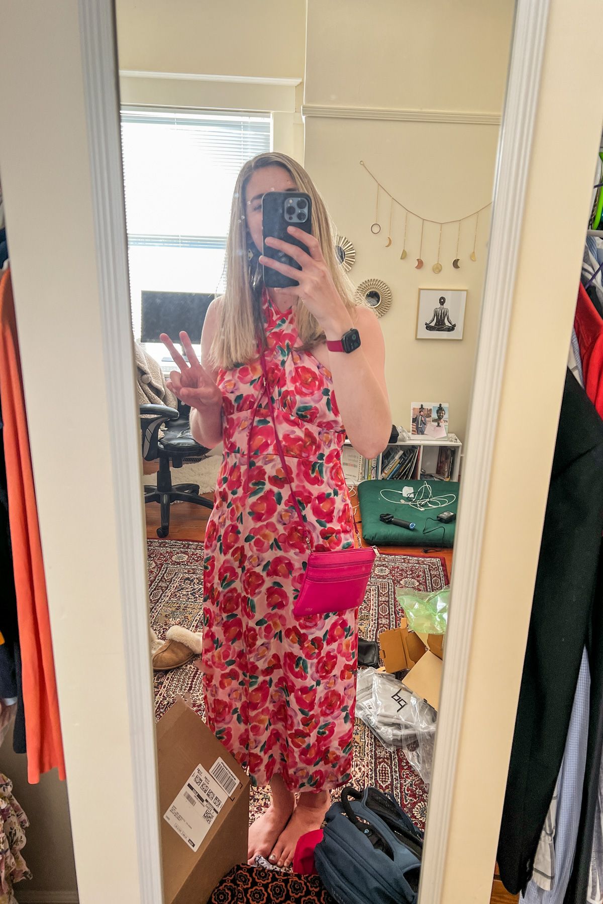 A woman wearing a silky, pink floral dress takes a mirror selfie with her bedroom visible in the background.