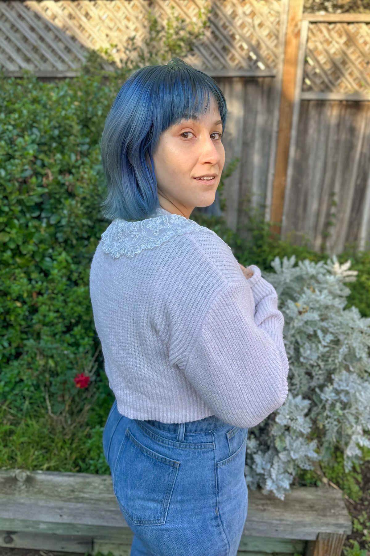 A blue-haired woman wearing a pink frilly cardigan and jeans looks back over her shoulder standing in front of a patch of grass in front of a backyard fence.