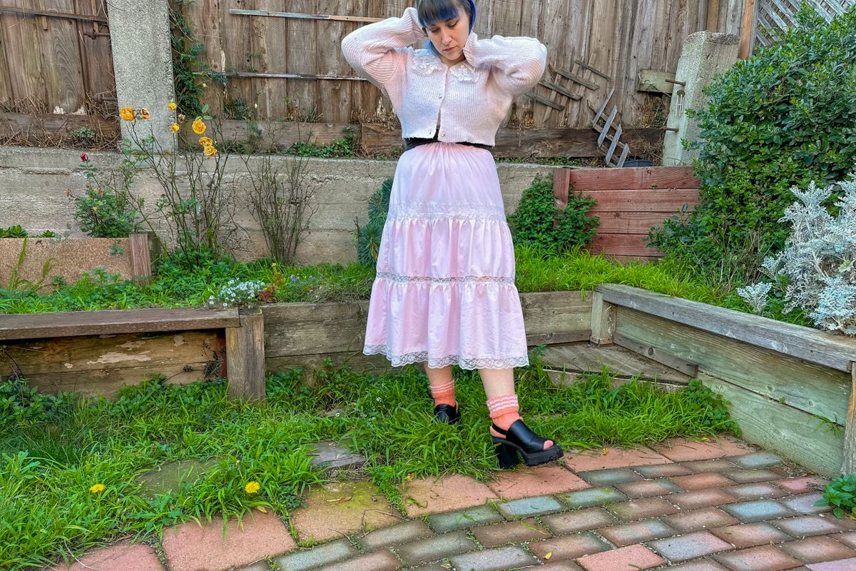 A blue-haired woman wearing a long, pink frilly skirt and matching cardigan, stands on a patch of grass in front of a backyard fence with her hands on her head.