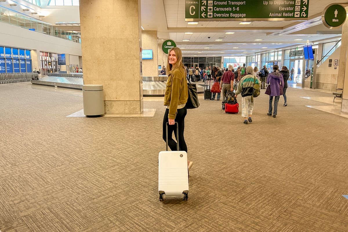 A woman wearing a brown jacket and black leggings looks back over her shoulder as she pulls a light blue suitcase through an airport baggage claim terminal.