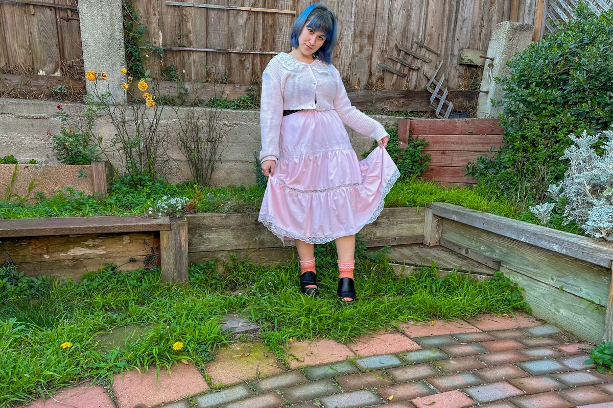A blue-haired woman wearing a long, pink frilly skirt and matching cardigan, stands on a patch of grass in front of a backyard fence, lifting her skirt in a curtsy.