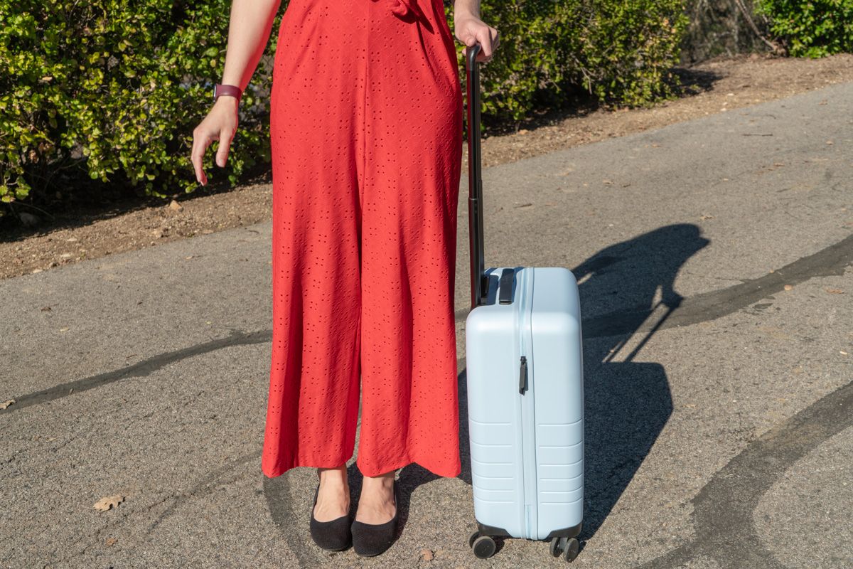 A woman's legs wearing a red jumpsuit and black flats stands next to a blue suitcase on a paved driveway on a sunny day.