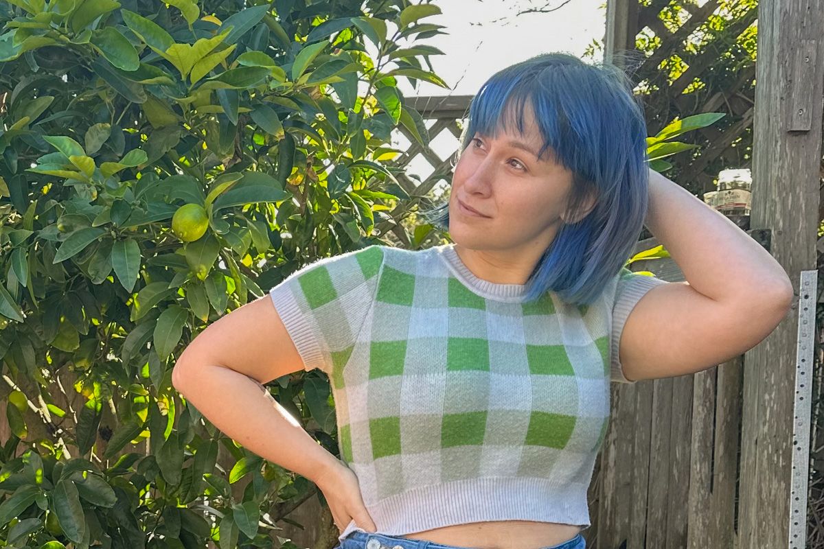 A blue-haired woman wearing a cropped, green and white checkered sweater scratches her head while standing next to a backyard lemon tree.