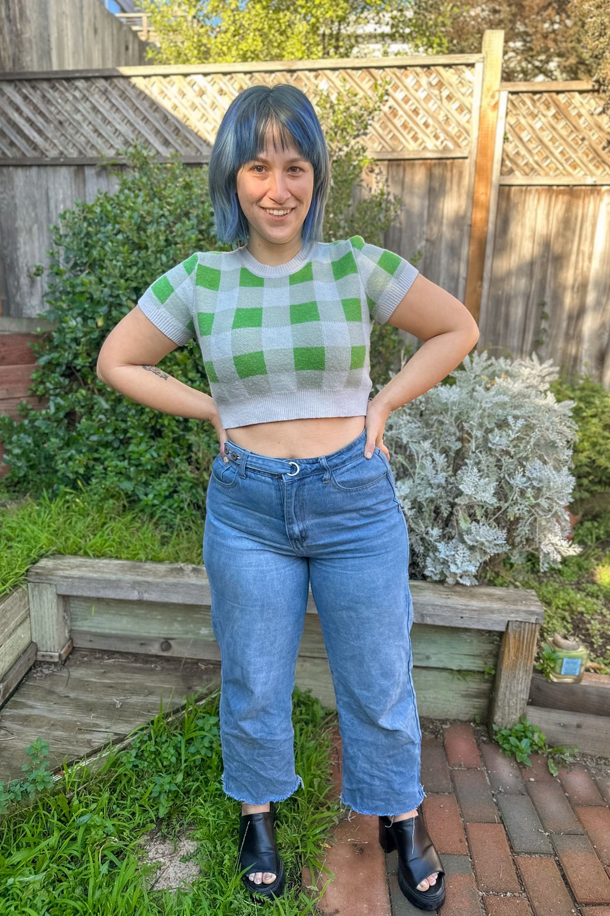 A blue-haired woman wearing a cropped, green and whiter checkered sweater and jeans stands with her hands on her hips on a brick patio next to a patch of grass in front of a backyard fence.