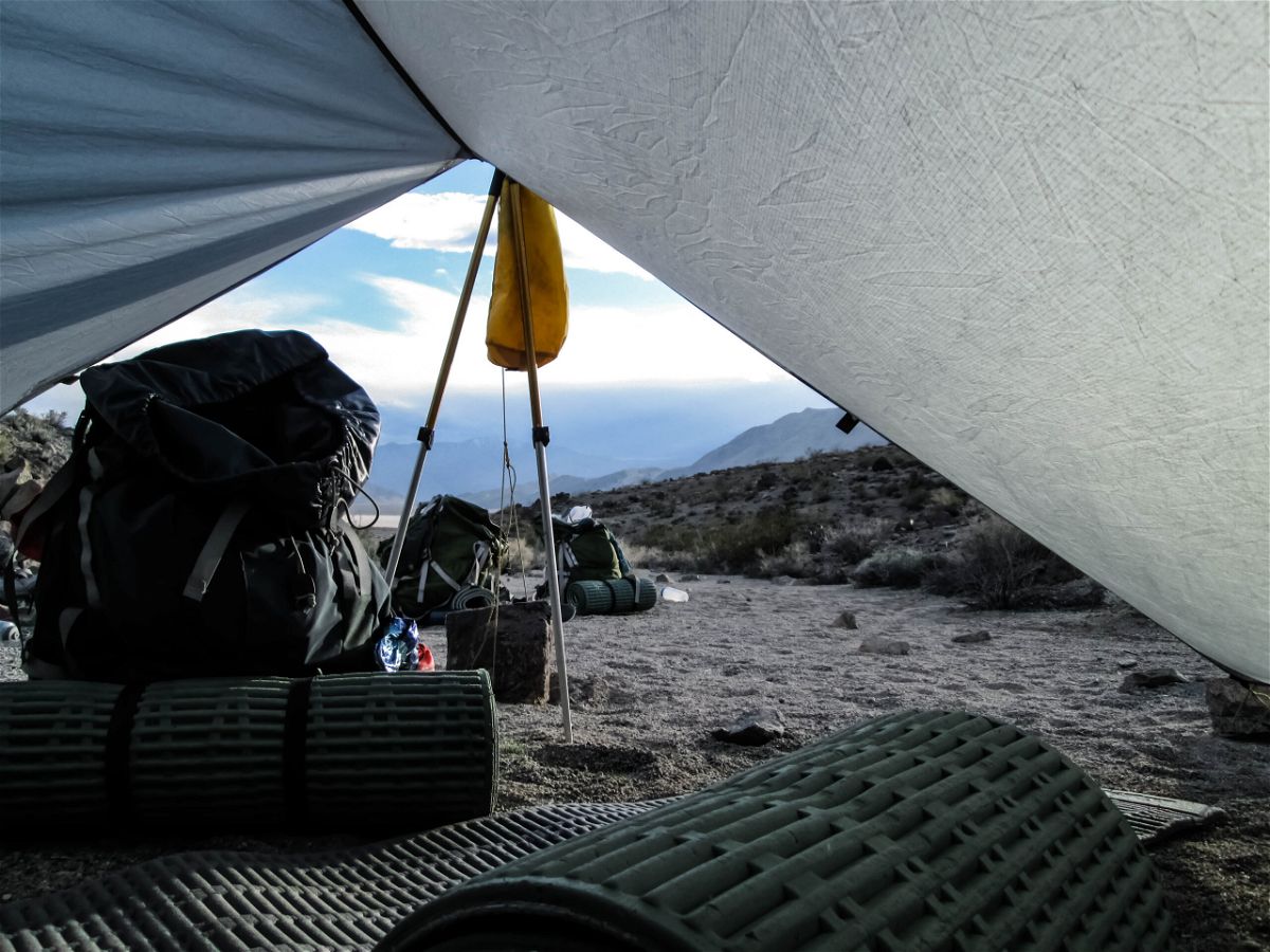 Looking out from inside of a tent, in the foreground are sleeping pads, a backpacking pack, and trekking poles, with more gear in the midground, and a rocky landscape in the background.