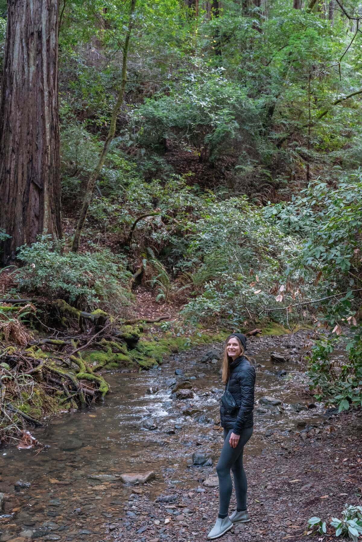 A woman in dark clothing standing beside a creek in Muir Woods, surrounded by towering redwoods.