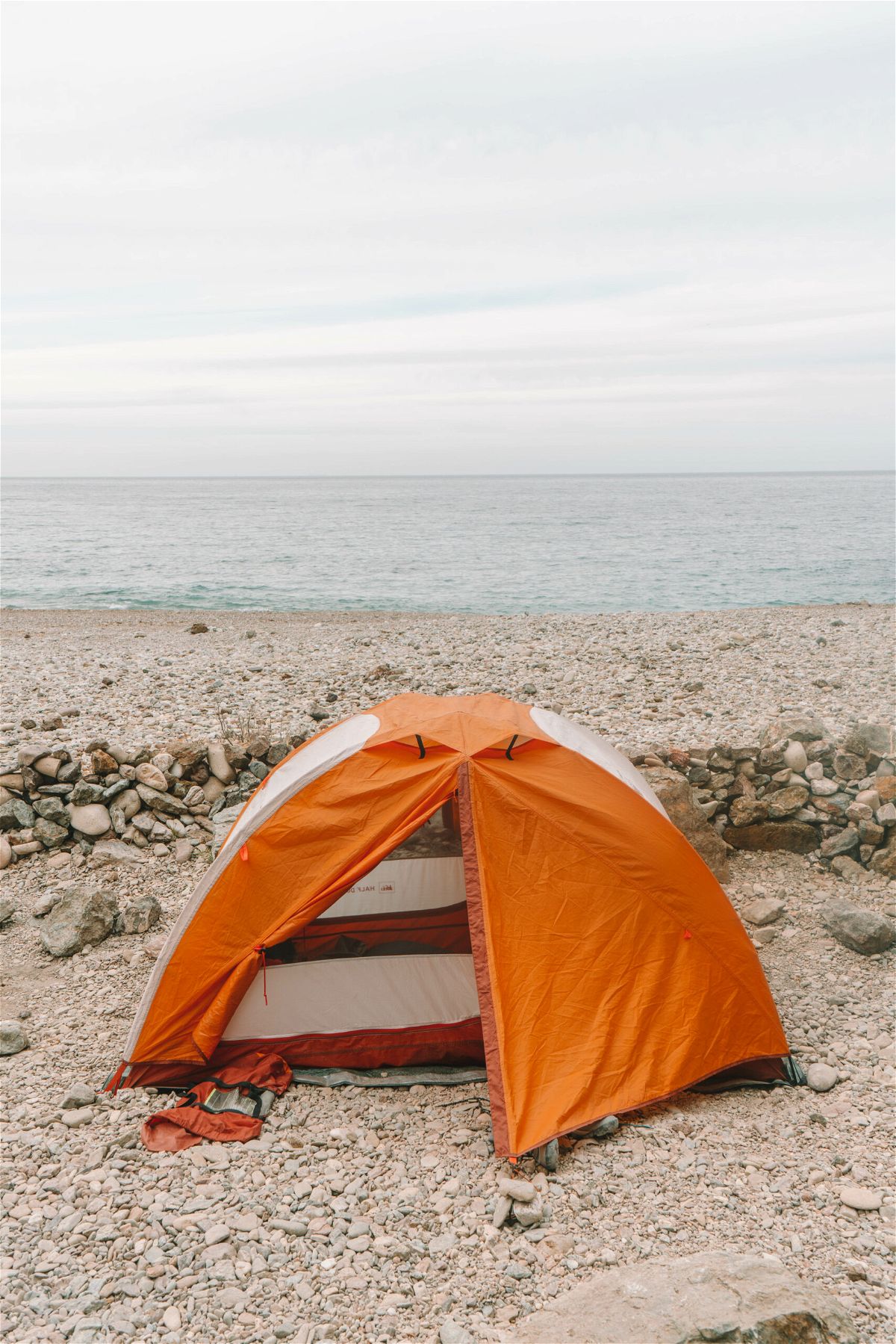 An orange and white dome tent set up on a rocky beach in front of the ocean.