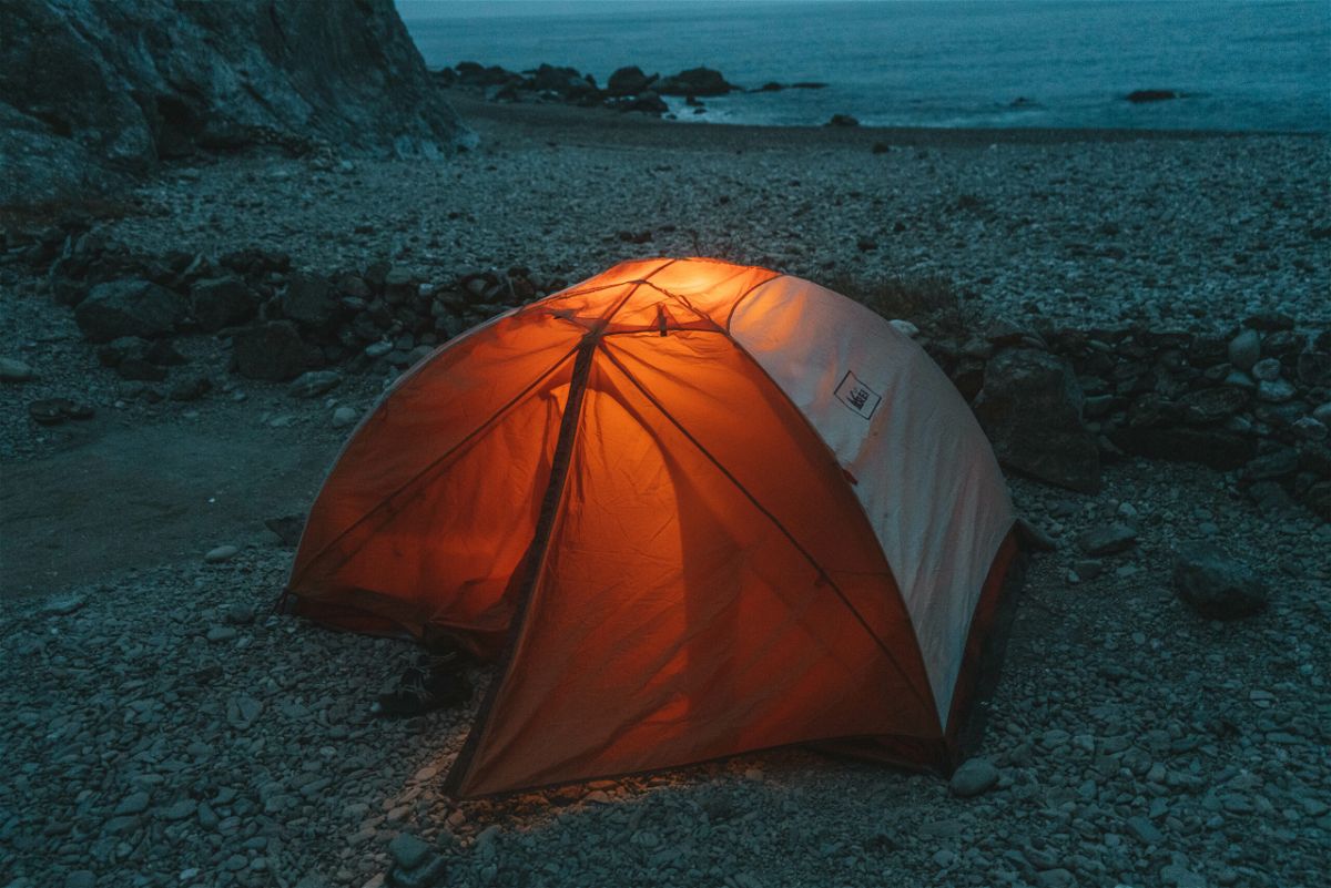 An orange and white tent with a lantern inside glows in the dark on a rocky beach with the ocean in the background.