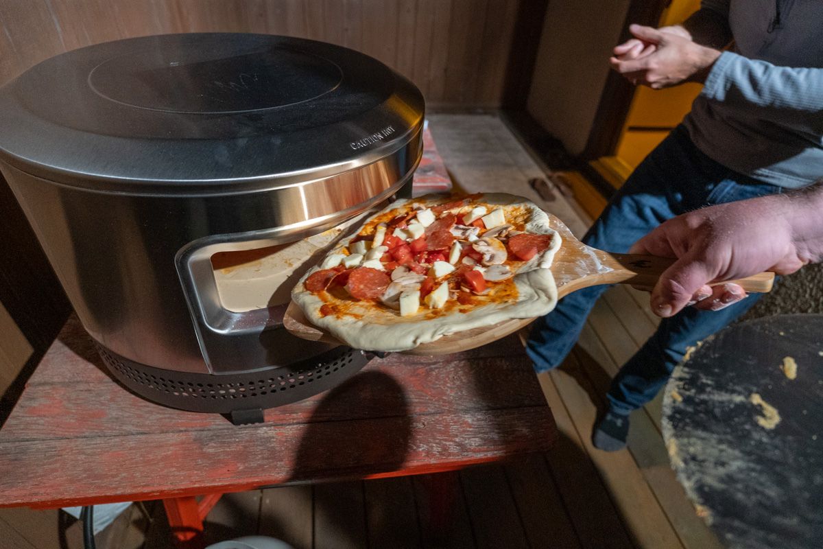 A hand holding a wooden paddle places a raw pizza inside of a Pi Prime Pizza Oven that sits on a wooden table in a backyard at dusk, with a brown fence behind it.
