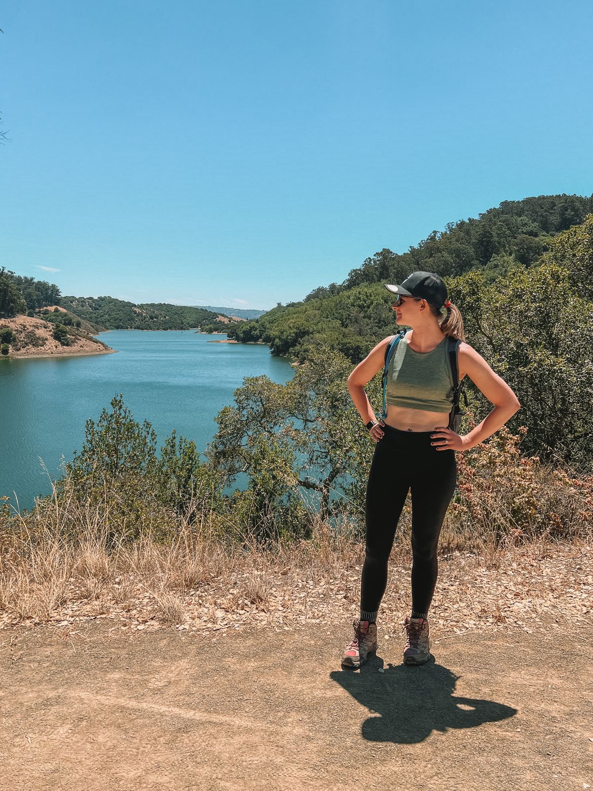 A woman hiker standing with her hands on her hips, looking at the lake to her right.