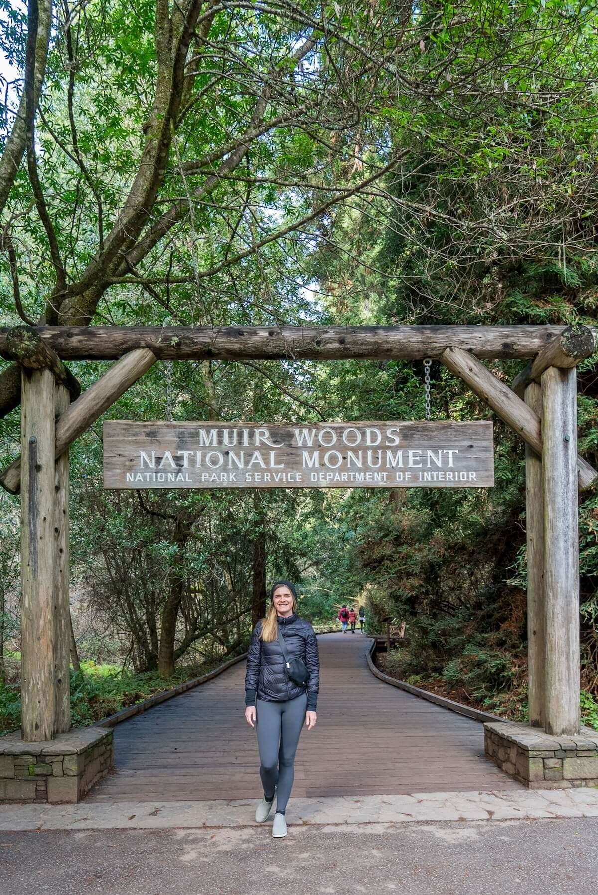 A female hiker in grey leggings and a black windbreak standing beneath a wooden sign marking the entrance to the Muir Woods Main Trail.