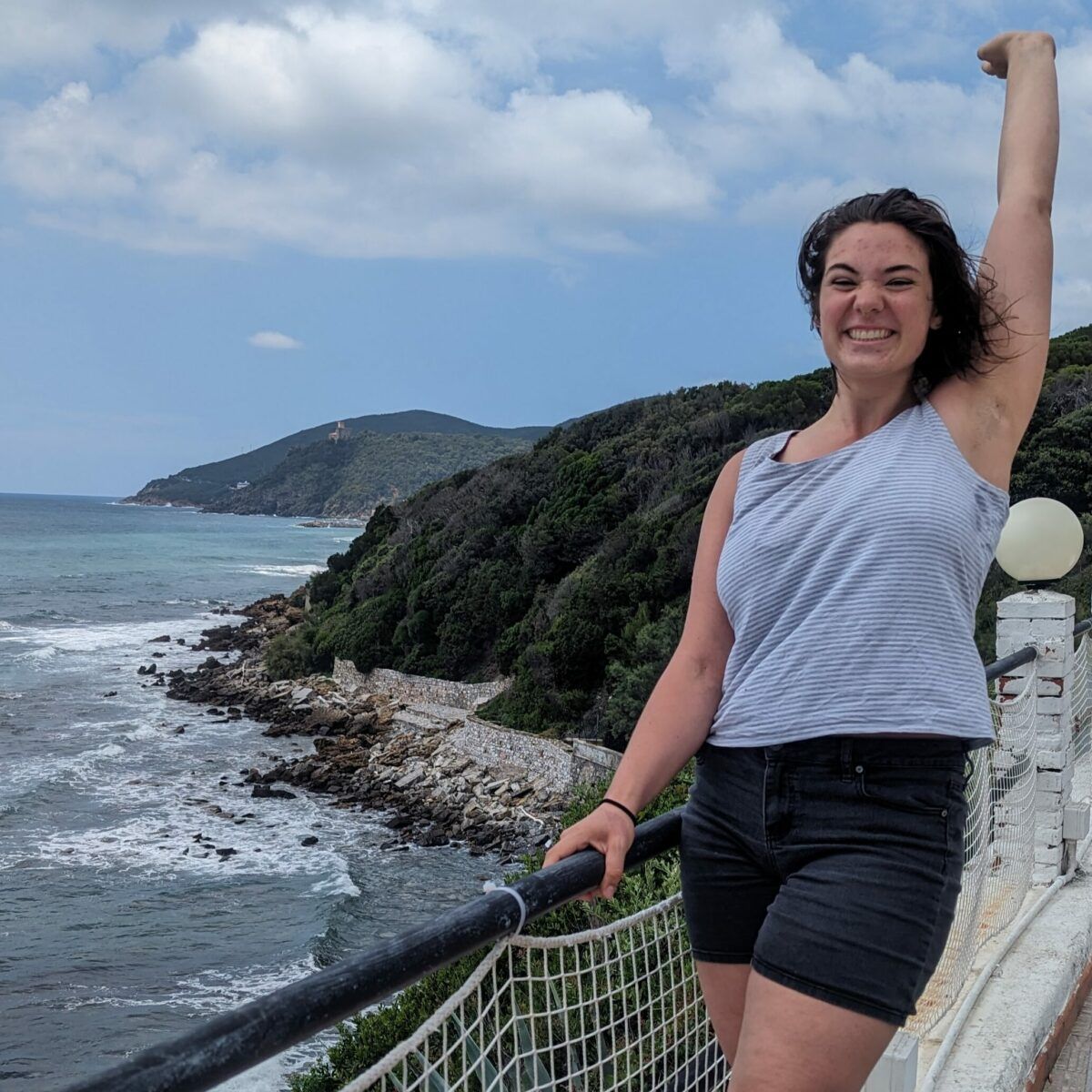 Jodelle posing at coastal lookout point, with one arm up in the air and the other hand holding the railing.