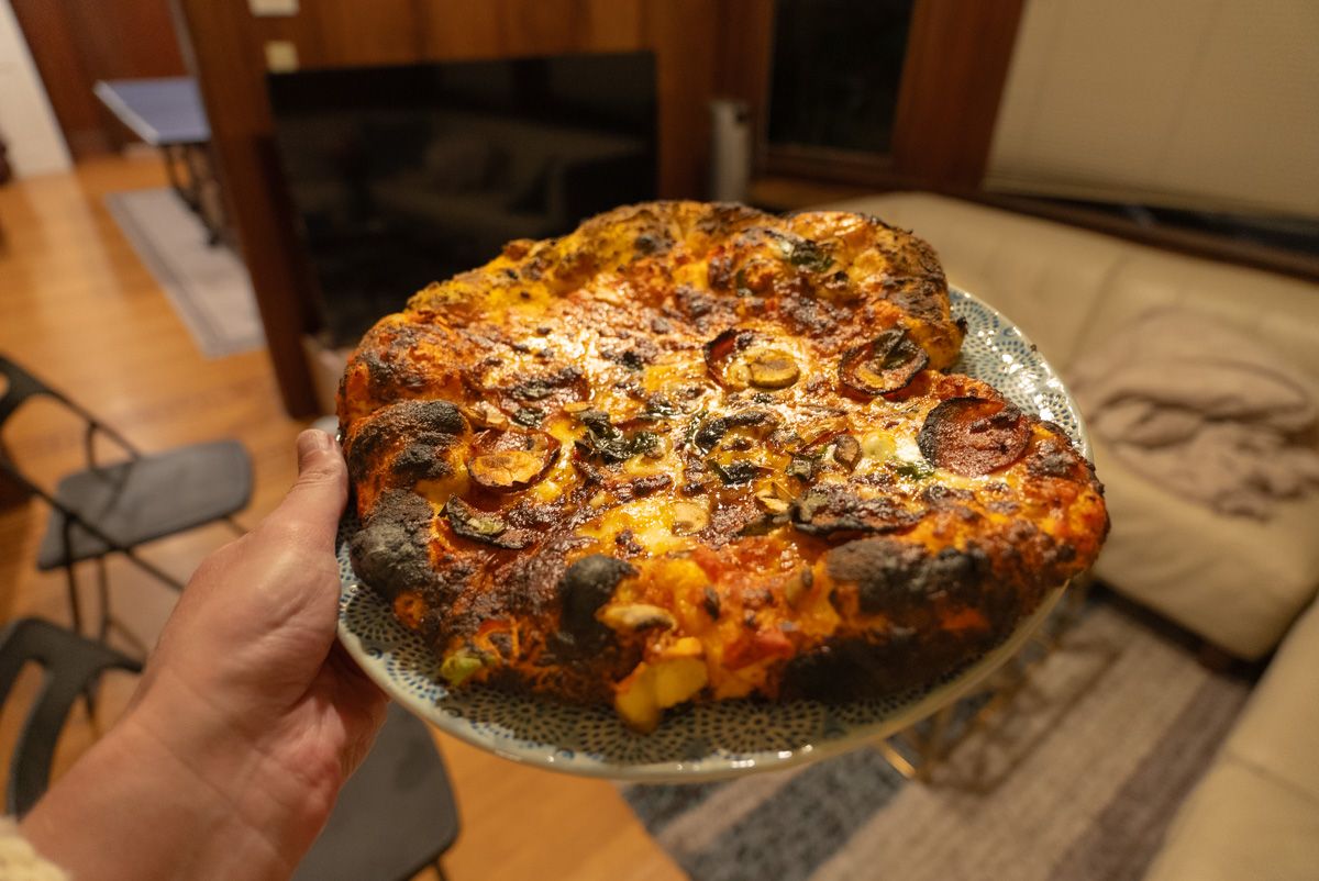 A hand holds a plate with a charred, toasty pizza sitting on it, and an interior living space in soft focus in the background.