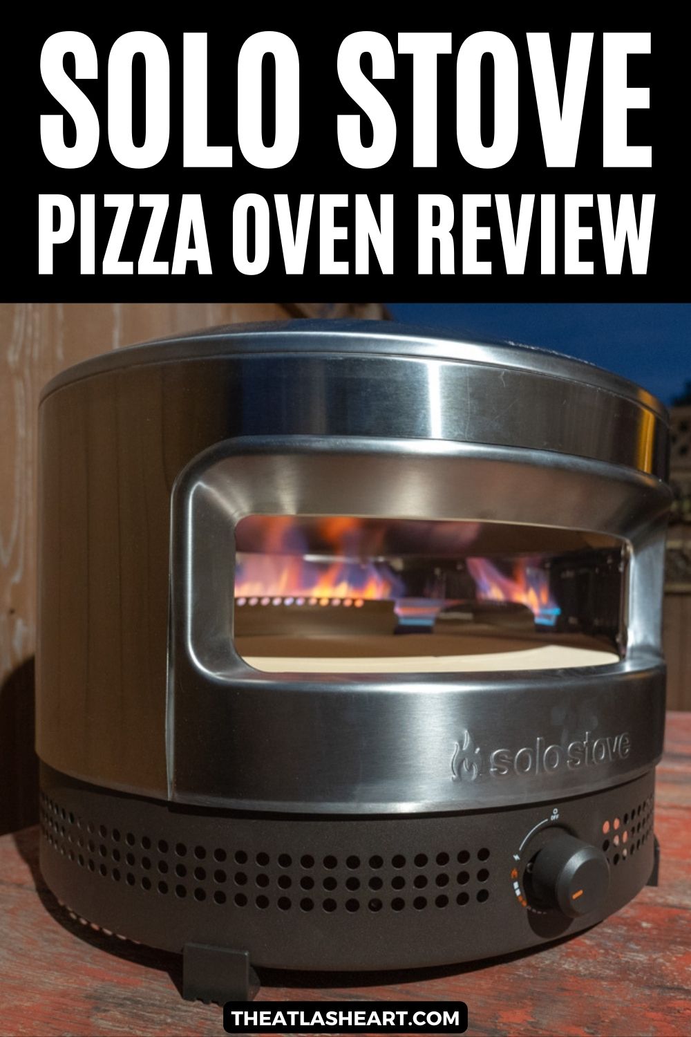 Flames visible inside a Pi Prime Pizza Oven sitting on a wooden table in a backyard at dusk, with a brown fence behind it, and the text overlay, "Solo Stove Pizza Oven Review."