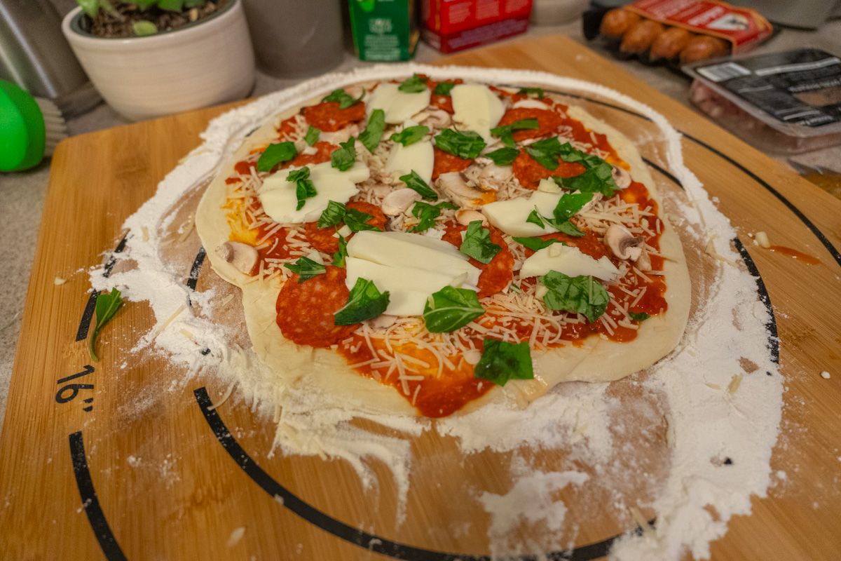 A raw pizza sitting on a floured wooden cutting board, loaded with chunks of mozzerella and fresh basil leaves. 