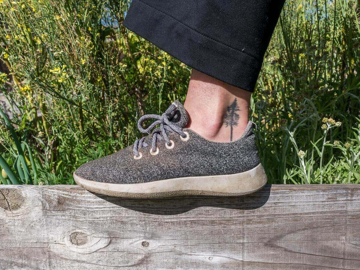 A foot wearing a grey sneaker propped up on a weathered wooden ledge with wildflowers behind it.