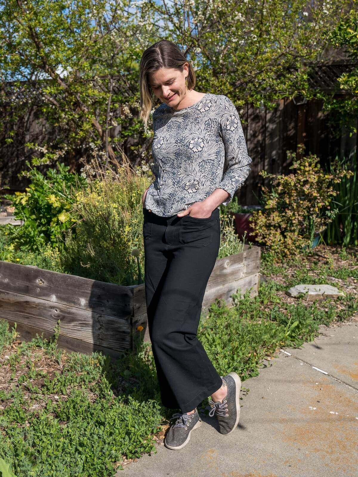A young woman in black pants, a grey sweater, and grey sneakers poses on a sidewalk with a green front garden behind her.