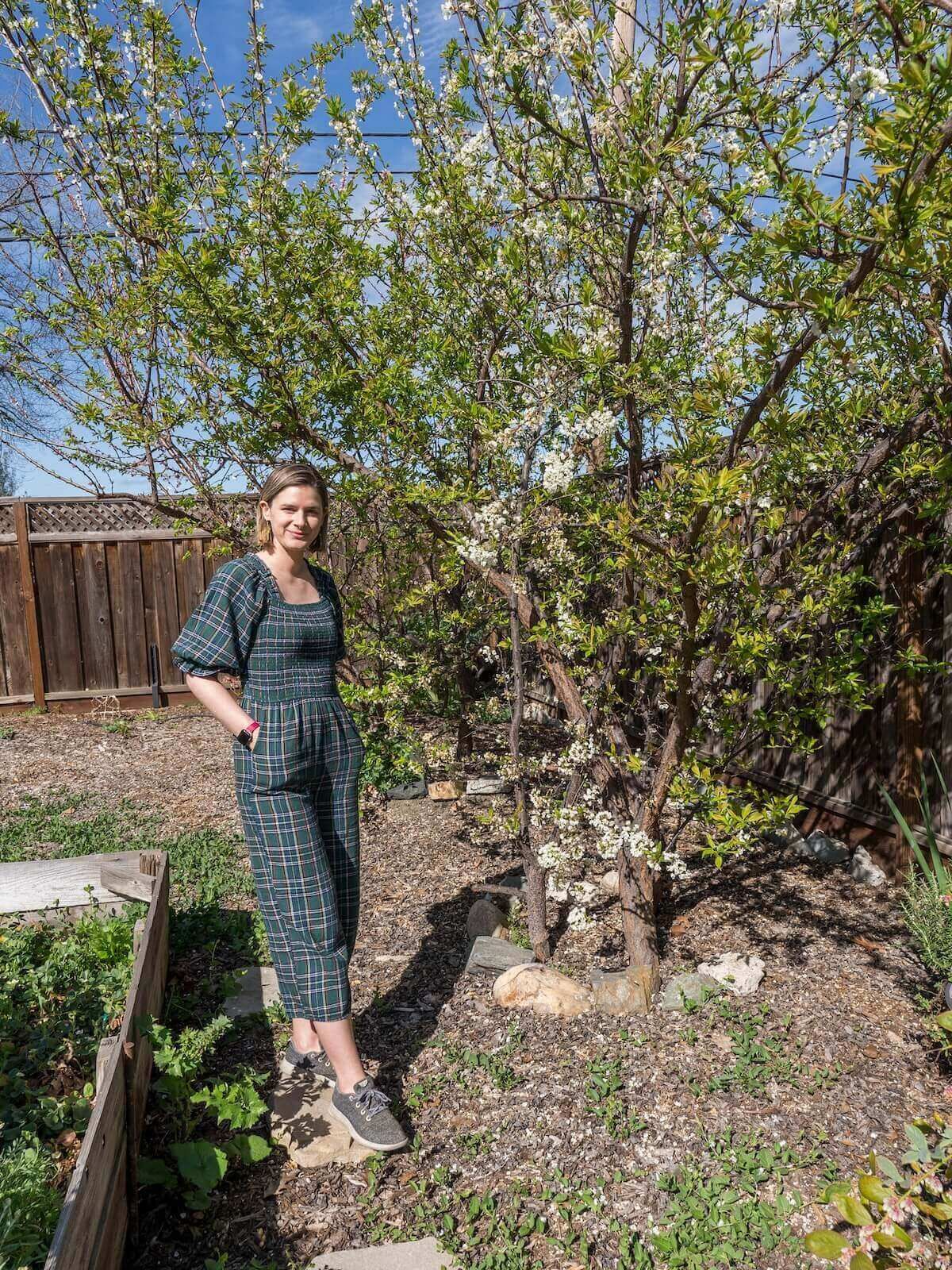 A young woman wearing a plaid jumpsuit and grey sneakers poses next to a planter box in a green garden, with a tree behind her.