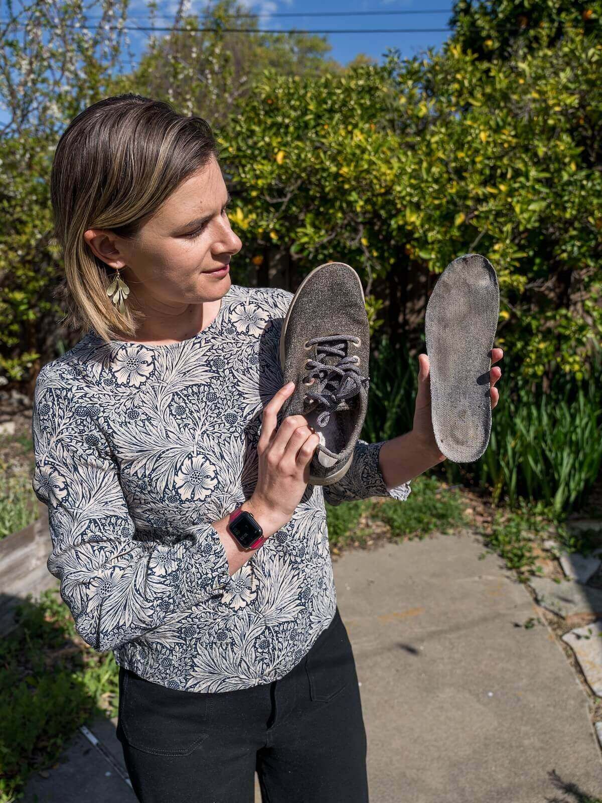 A woman in a grey floral-patterned shirt holds up a grey sneaker and its insole with a lush garden behind her.
