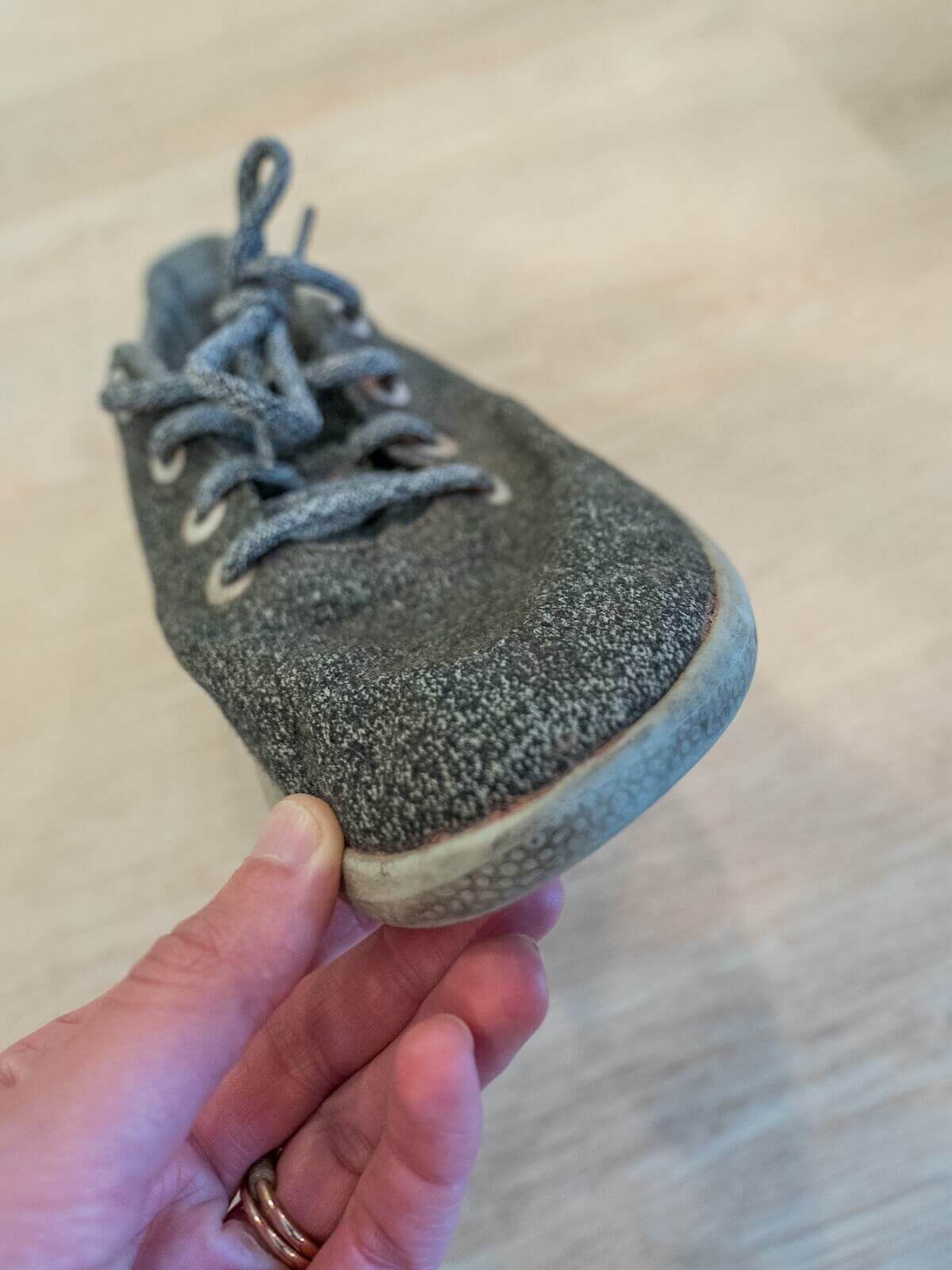 A hand holds the toe of a somewhat worn grey sneaker with a light hardwood floor behind it.