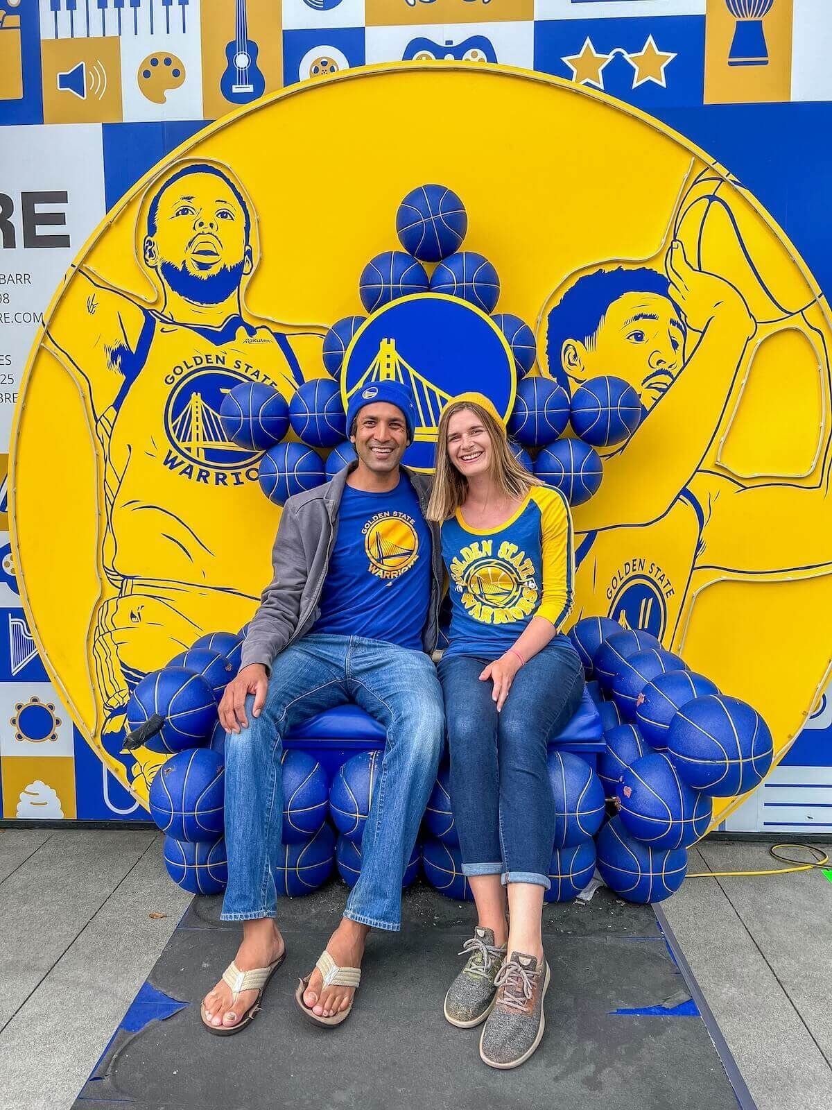 A man and a woman deck out i a yellow and blue Warriors gear poses in front of a Warriors backdrop.