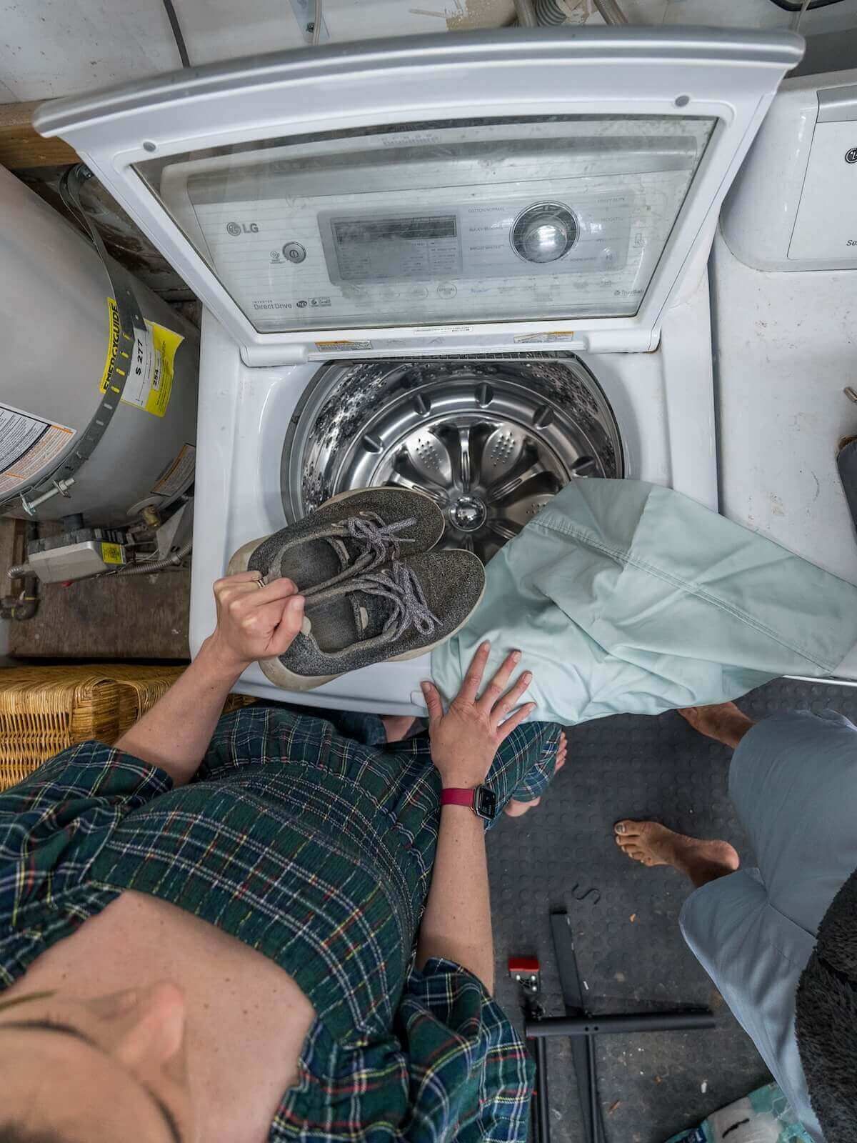 A view from above of a young woman wearing dark green plaid preparing to place a pair of grey sneakers into a washing machine.
