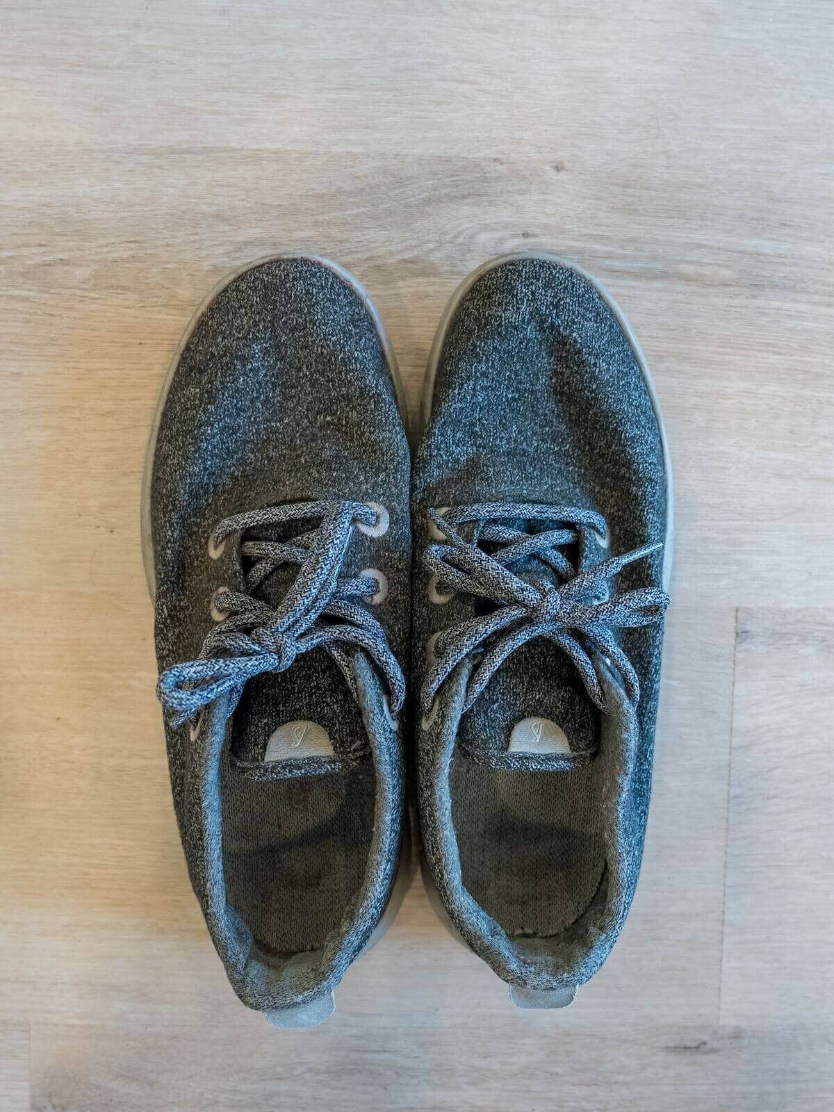A pair of somewhat worn grey sneakers seen from above, sitting on a light hardwood floor.