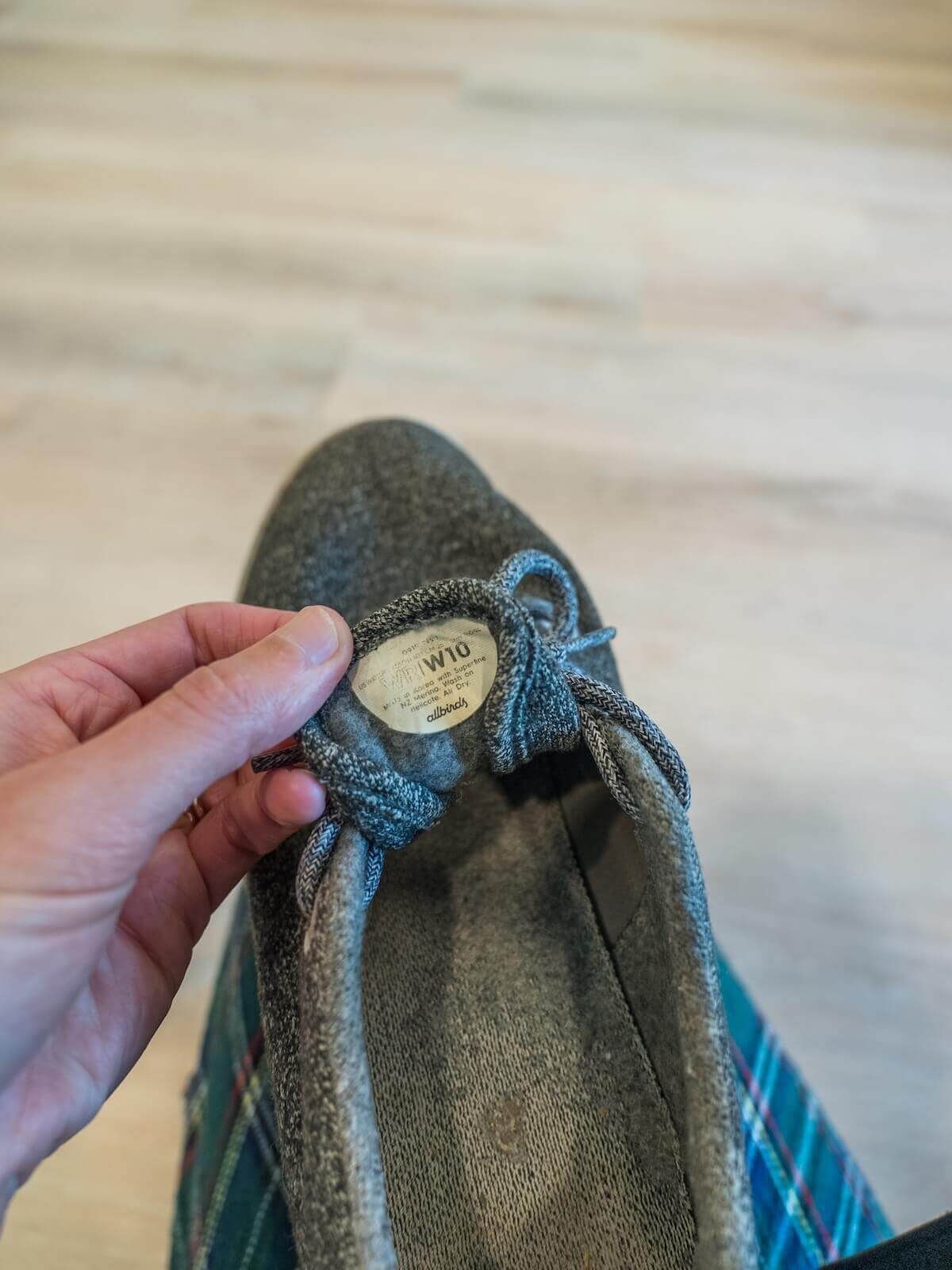A hand pulls out the tongue of a grey sneaker to show the label, with a light hardwood floor behind it.