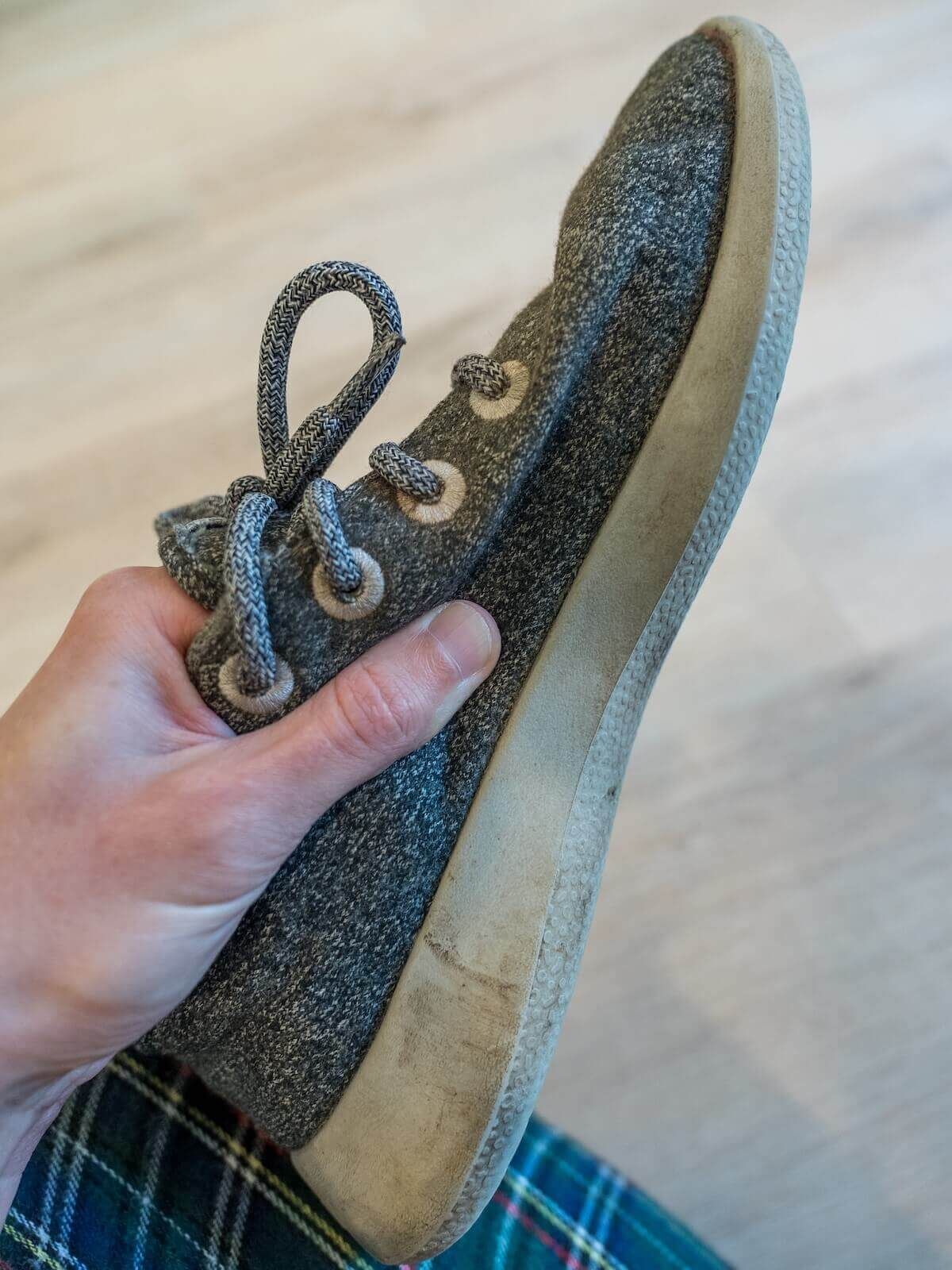 A hand holds a somewhat worn grey sneaker with a light hardwood floor behind it.