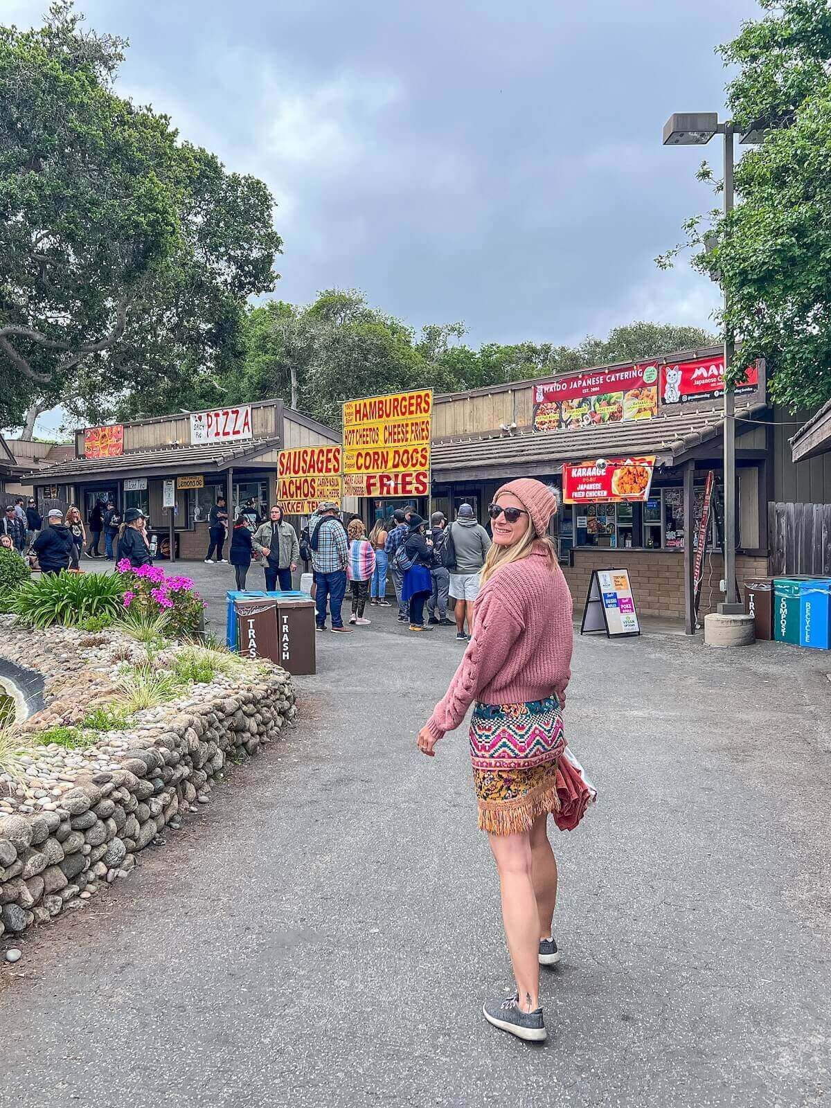 A young woman in a pink sweater and patterned miniskirt wears grey sneakers and smiles back over her shoulder while walking on a path towards some outdoor food stands.