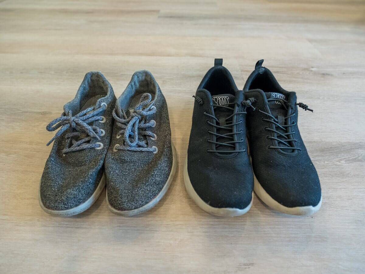 A pair of slightly worn grey sneakers sitting next to a pair of black sneakers on a light hardwood floor.
