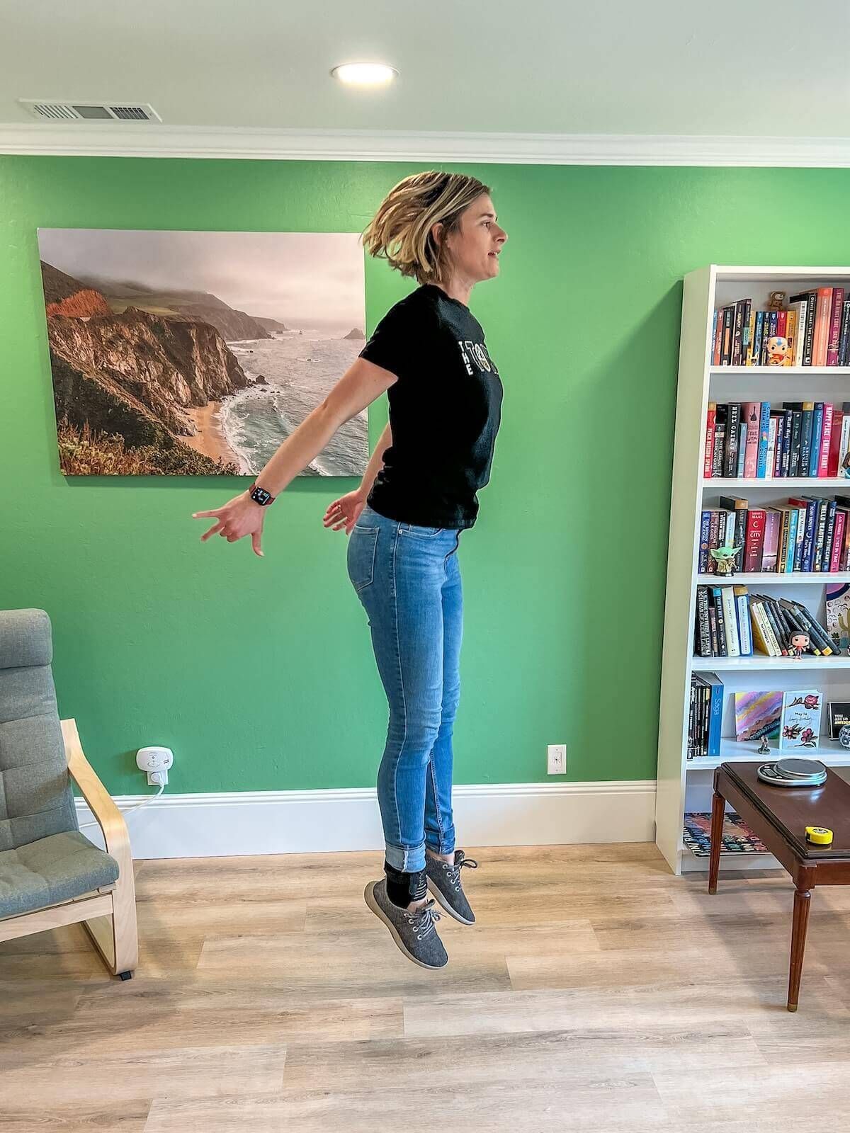 A light-haired young woman wearing jeans, a black t-shirt, and grey sneakers caught mid-jump in an interior with a green wall behind her.