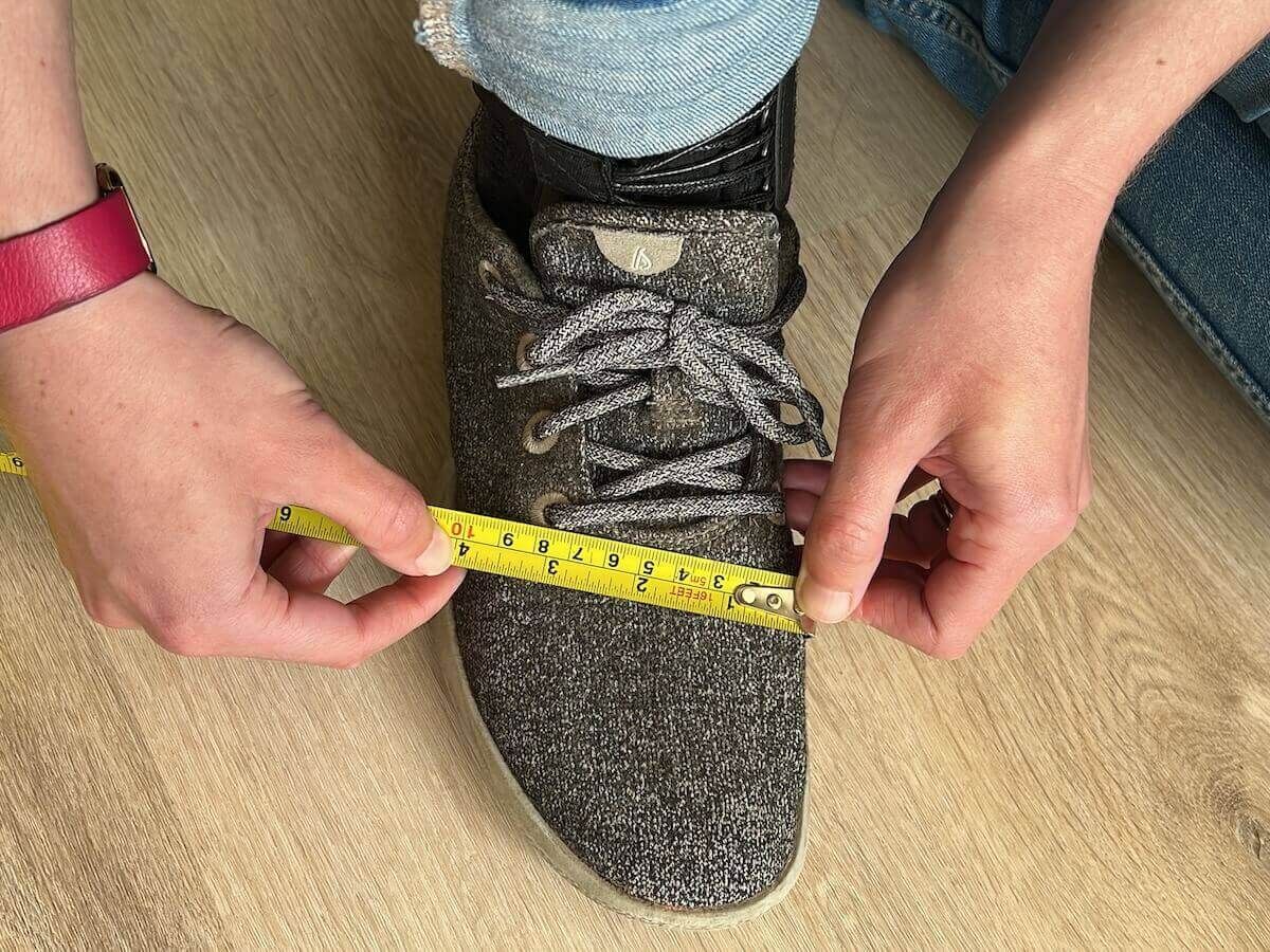 A pair of hands measuring the width of a foot wearing a grey sneaker against a hardwood floor.