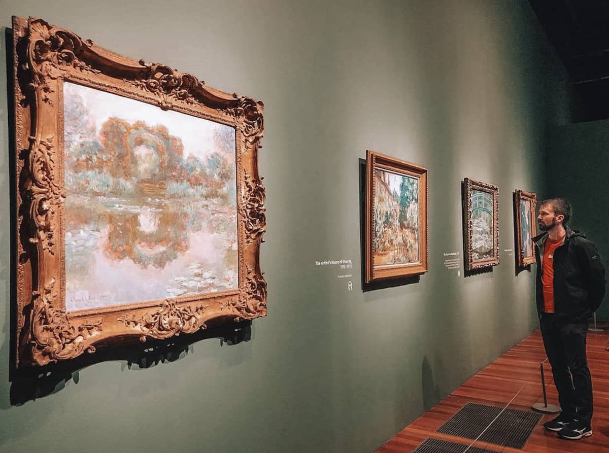 A man looks at impressionist paintings in gold frames hanging on a sage green museum wall.