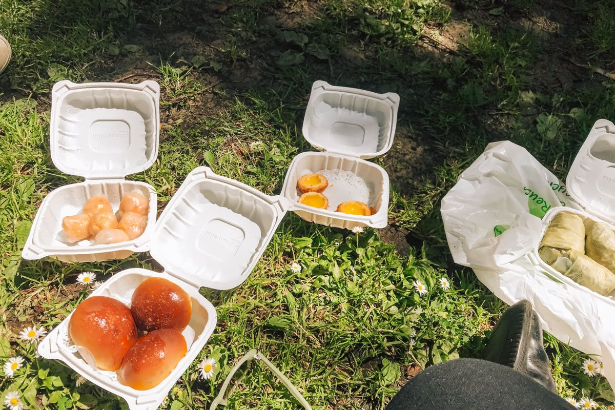 Four white plastic to-go containers full of dim-dum on white doilies sit open on daisy-studded grass.