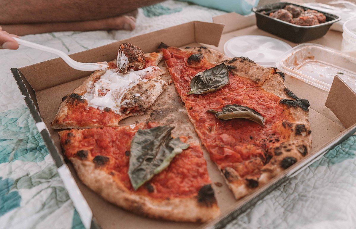 A close-up of a gourmet pizza in a cardboard box sitting on a picnic blanket.