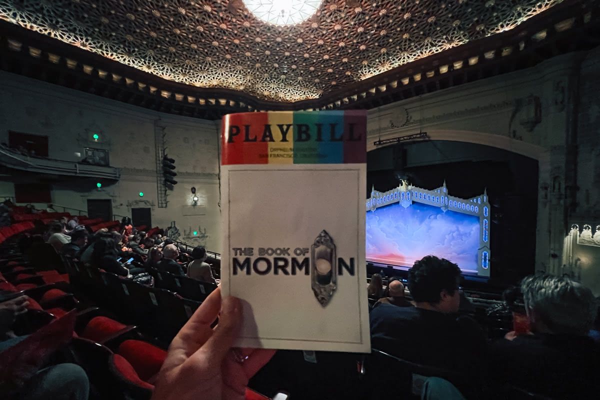 A hand holds up a playbill for 'Book of Mormon' in a crowded theatre with the stage visible beyond.
