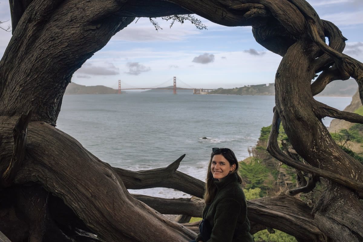 A young woman in a dark fleece pullover smiles over her should in front of a circular formation of wild-blown trees that frame a view of the ocean and the Golden Gate Bridge.