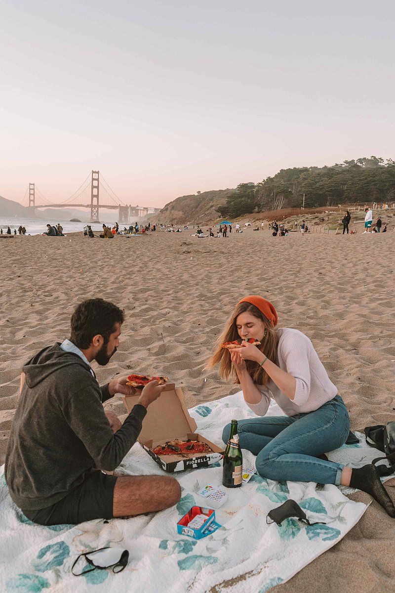 A man and woman sit across from each other on a picnic blanket on the beach, sharing a pizza, with the golden gate bridge in the background.
