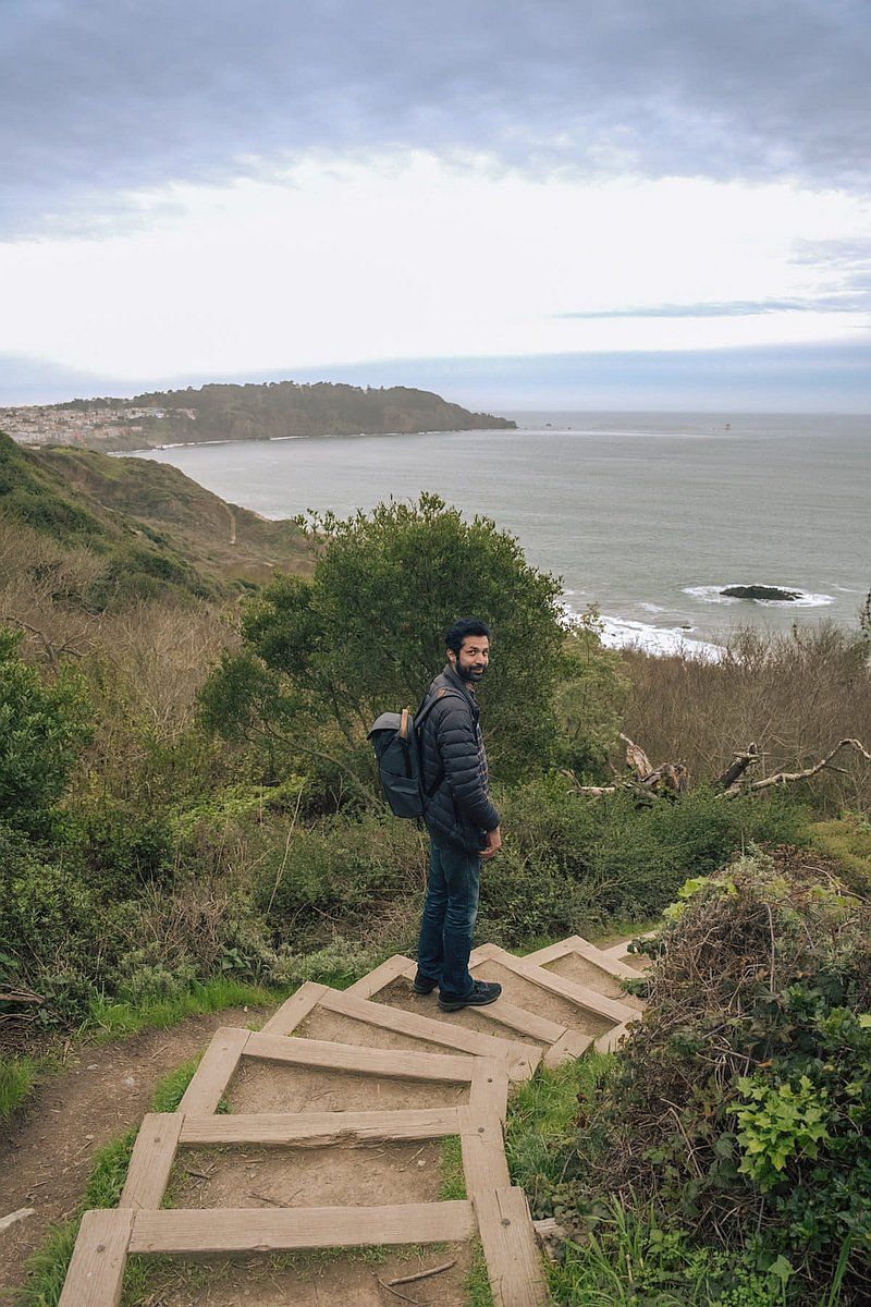 A man in a black down jacket and a backpack looks back over his shoulder while standing on a set of wooden stairs that descend towards a beach.