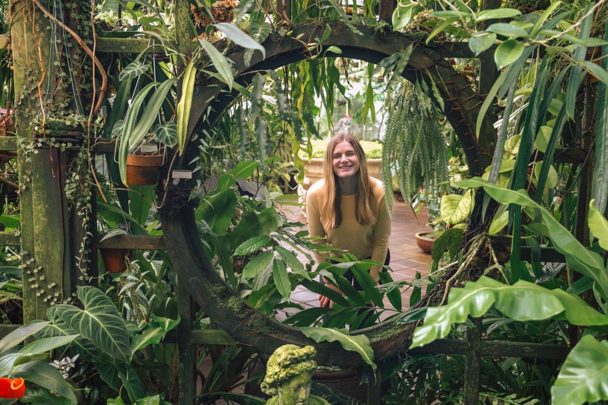 A blonde woman in a yellow sweater smiles as she peers through a circular opening in a wall of lush garden greenery.