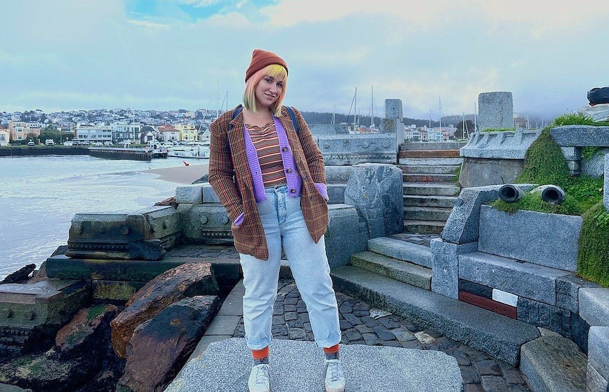 A woman with orange and yellow hair wearing a brown plaid blazer, burnt orange beanie, and jeans smiles at the camera as she stands on sculptural rocks with a harbor and seaside cityscape in the background.