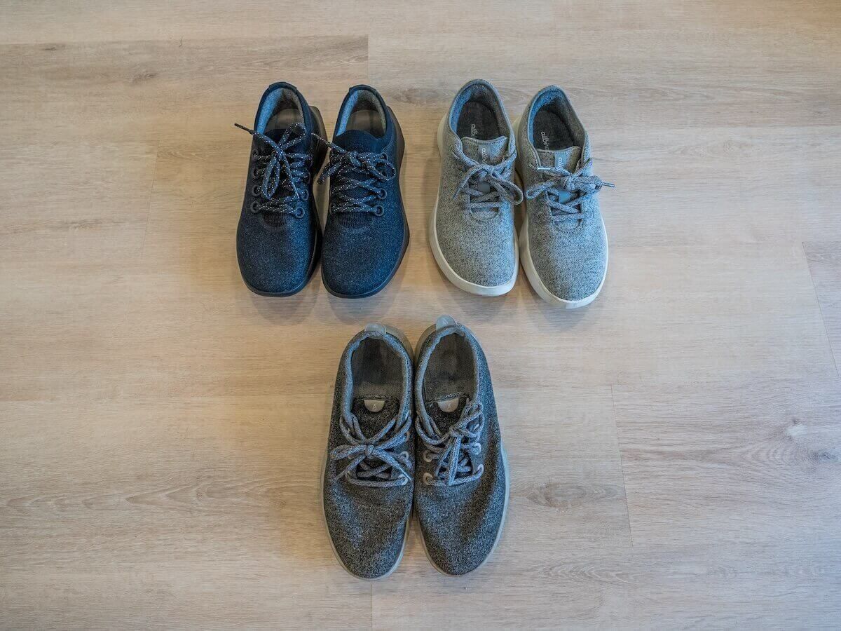 Three pairs of sneakers, black, light grey, and dark grey, sitting on a light hardwood floor.