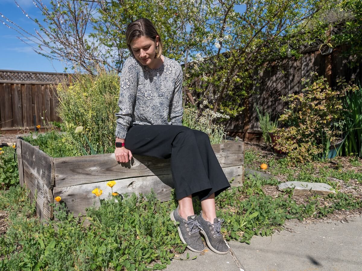 A woman in a grey floral-patterned shirt, black pants, and grey sneakers sits on the edge of a planter box with a lush garden behind her.