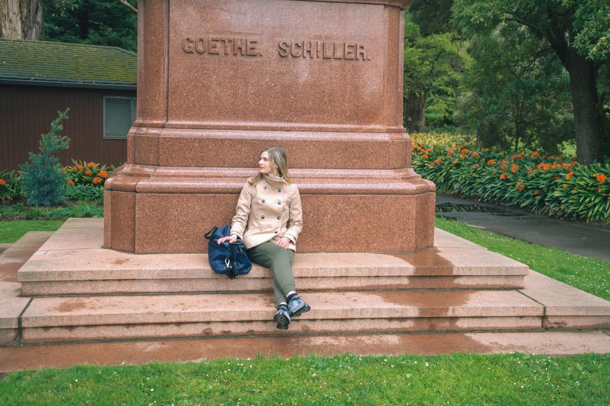 A young, light-haired woman in a tan trenchcoat sits against a brown concrete base of a public sculpture in a city park.
