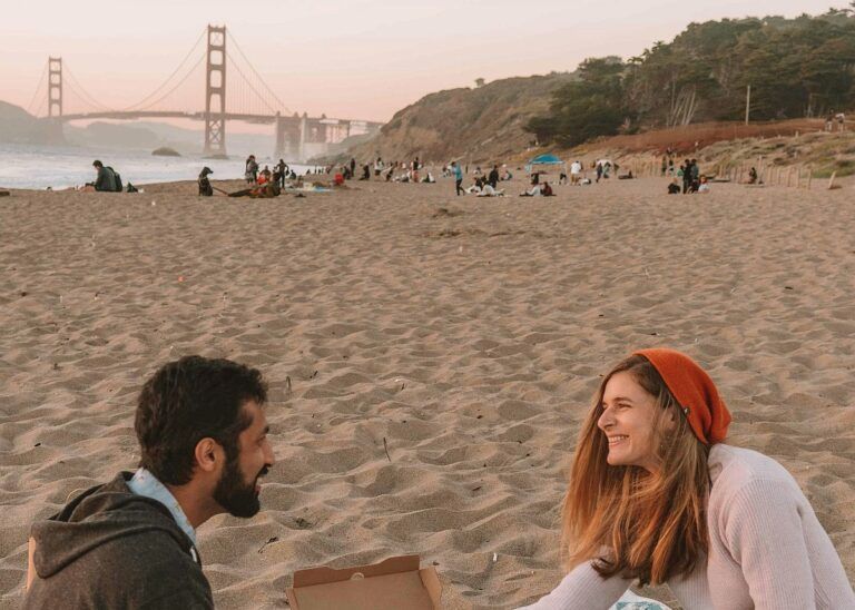 A man and a woman smile at each other as they sit on a beach at sunset, the golden gate bridge visible behind them, on one of the top date ideas in San Francisco.