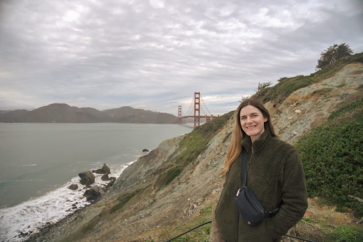 A young woman in a dark green fleece poses on a windswept beach with the Golden Gate Bridge in the background.