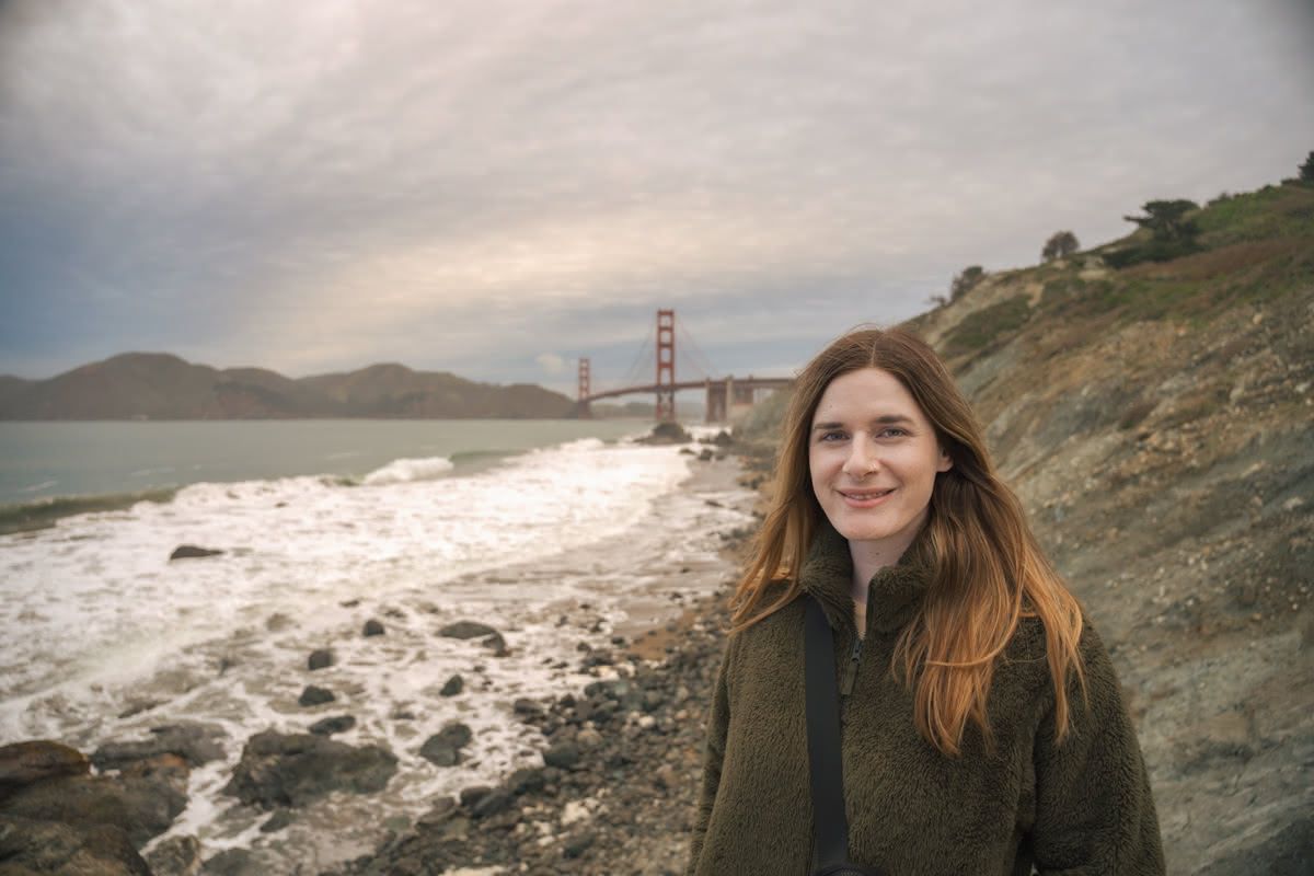 A young woman in a dark green fleece poses on a windswept beach with the Golden Gate Bridge in the background.