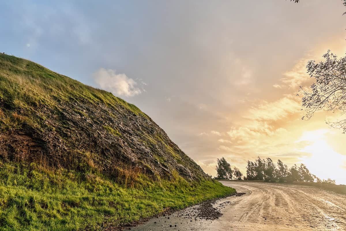A curving, wet road lined by a rocky slope covered in green grass with a golden sunset beyond.